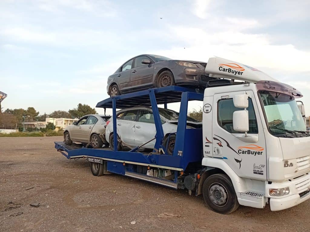 Truck-mounted car carrier carrying a dark gray sedan on a blue trailer, with 'CarBuyer' branding on the truck cab.