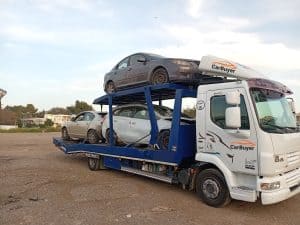 Truck-mounted car carrier carrying a dark gray sedan on a blue trailer, with 'CarBuyer' branding on the truck cab.